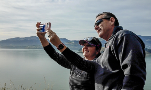 A man and women taking selfie with habour and hills in the background.